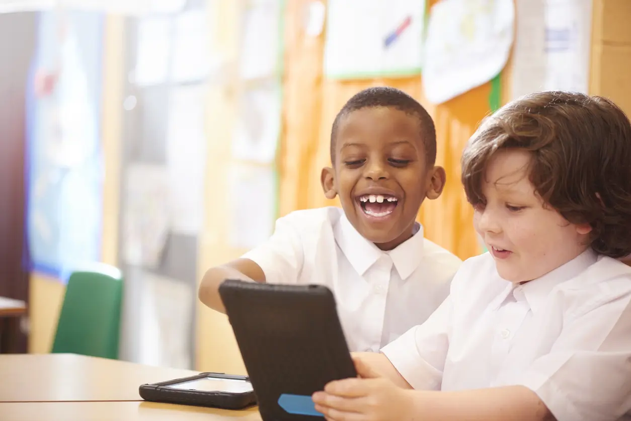 two primary aged schoolchildren sit at their desks and look engrossed in the work that they are doing on their digital tablets. The little boy has an amazed expression whilst looking at his tablet  and having fun . They are all wearing white school shirts