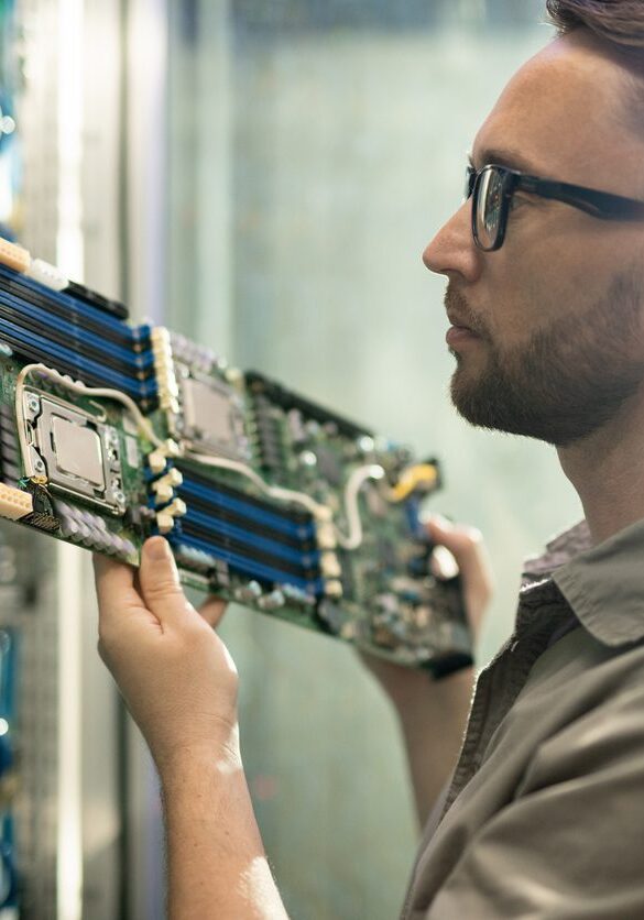 Server installation specialist working in datacenter room Serious skilled young bearded server installation specialist in glasses holding circuit board and examining server racks while working in datacenter room